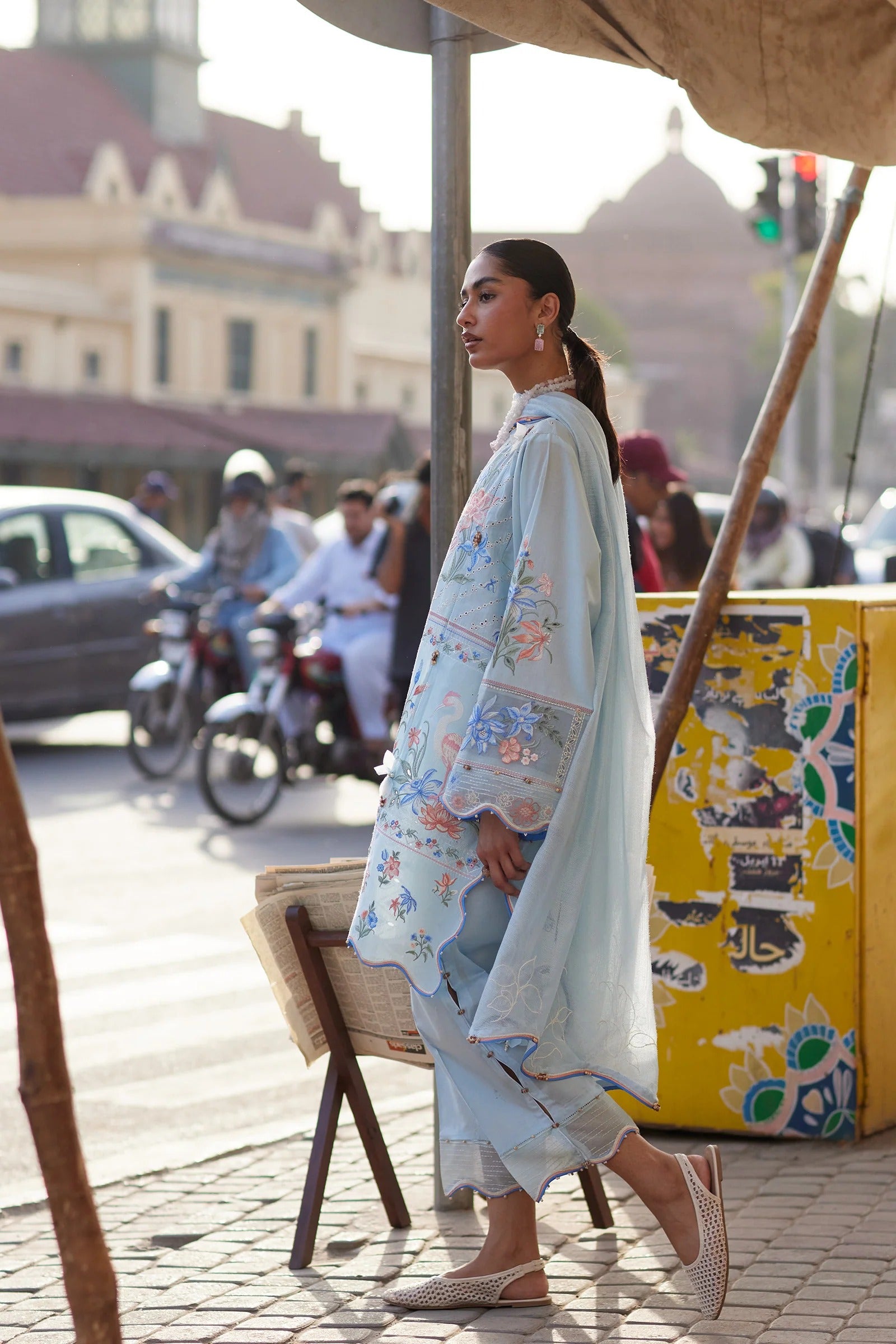 Woman in a light blue embroidered outfit walking on a street with buildings and people in the background.