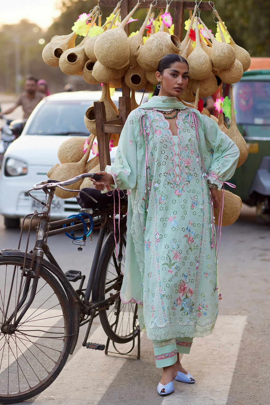 Woman in a light green traditional outfit standing next to a bicycle with woven baskets on a street.