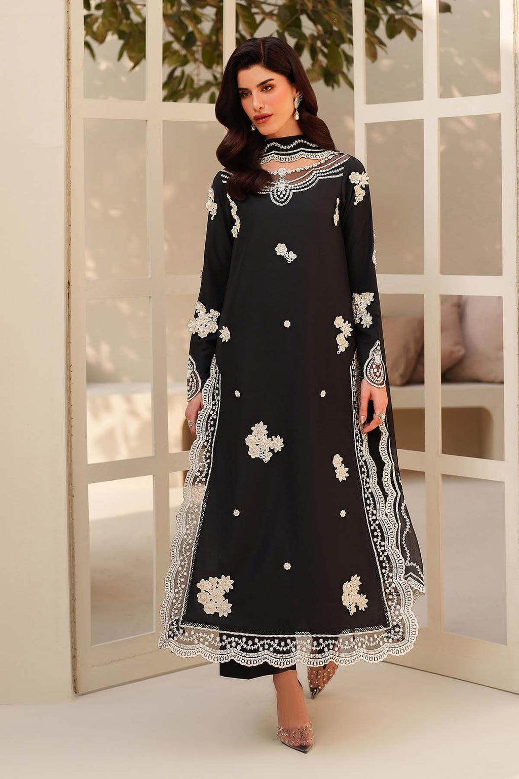 Woman wearing a black dress with white floral embroidery in a room with large windows.