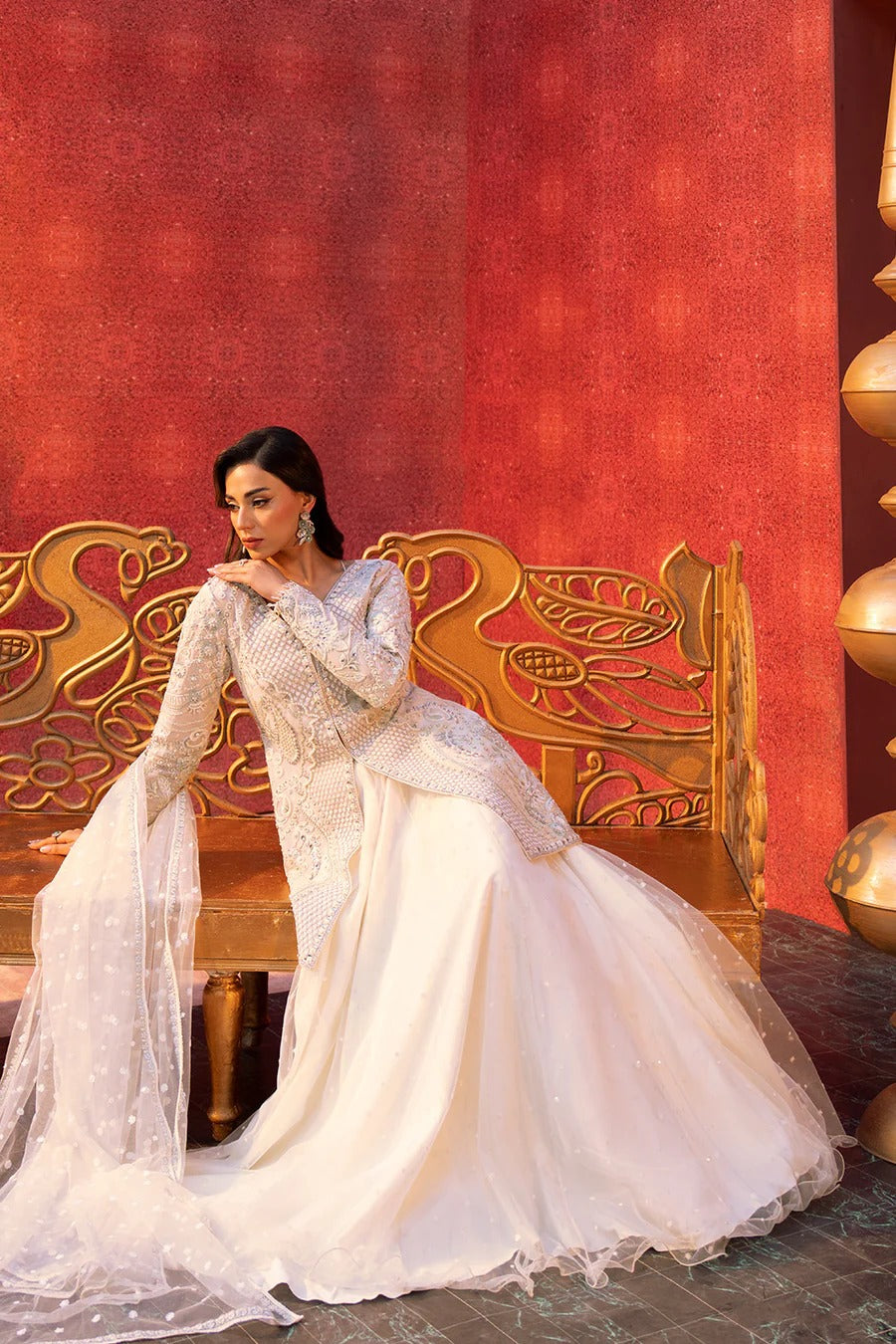 Woman in a white traditional outfit sitting on an ornate chair against a red wall.