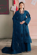 Woman wearing a blue embroidered outfit in a room with pink walls and decorative items.
