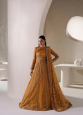 A woman stands in a minimalist cream-colored architectural setting wearing a heavily embroidered mustard yellow traditional gown with a matching sheer dupatta.