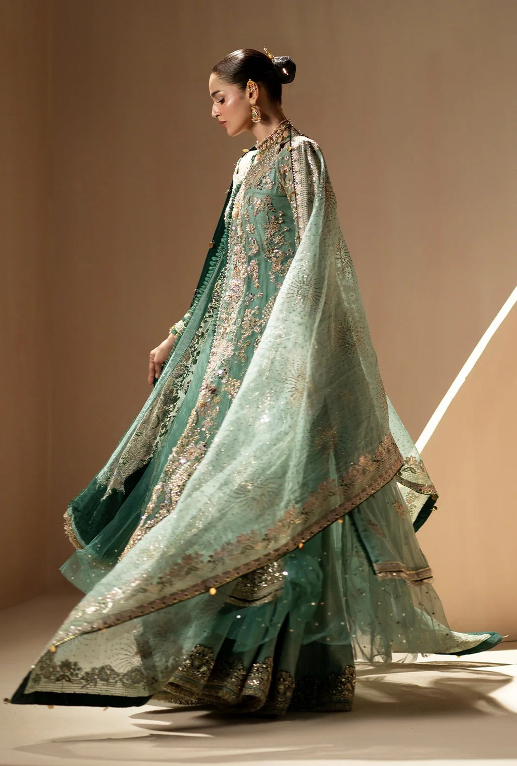 A model wearing a traditional green embroidered ethnic outfit with a matching sheer dupatta, photographed in a studio setting against a warm beige background.