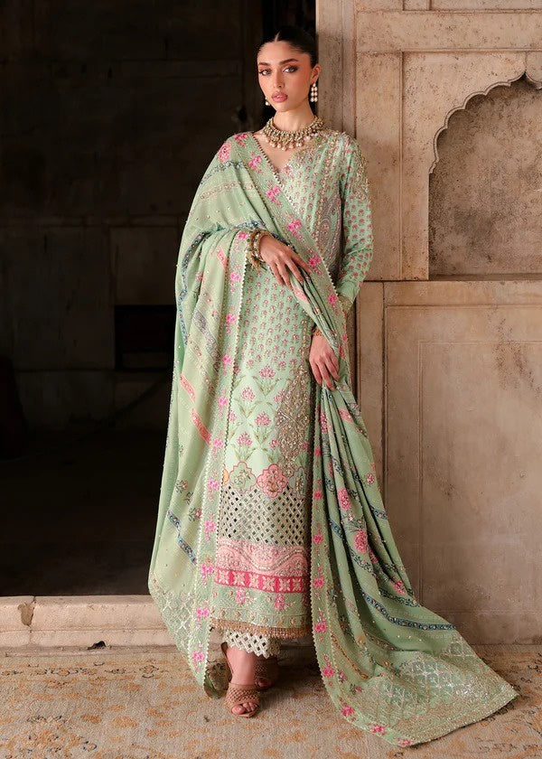 A woman models a sage green embroidered traditional outfit with pink floral accents and a matching dupatta, standing in front of a stone architectural background.