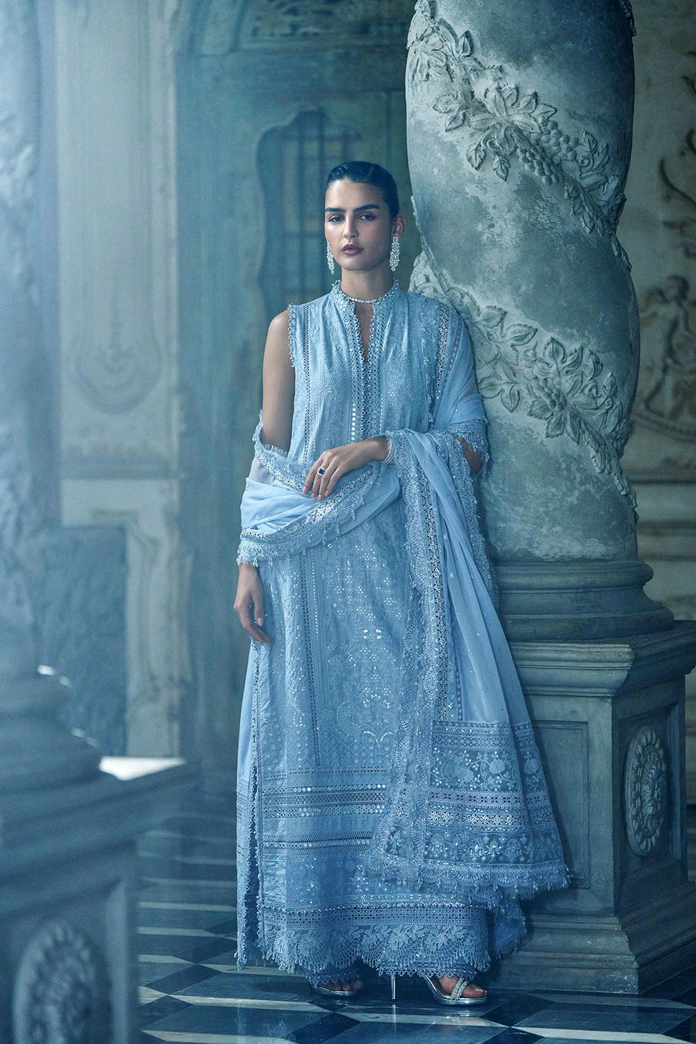 Woman in a light blue embroidered traditional outfit standing in an ornate architectural setting.