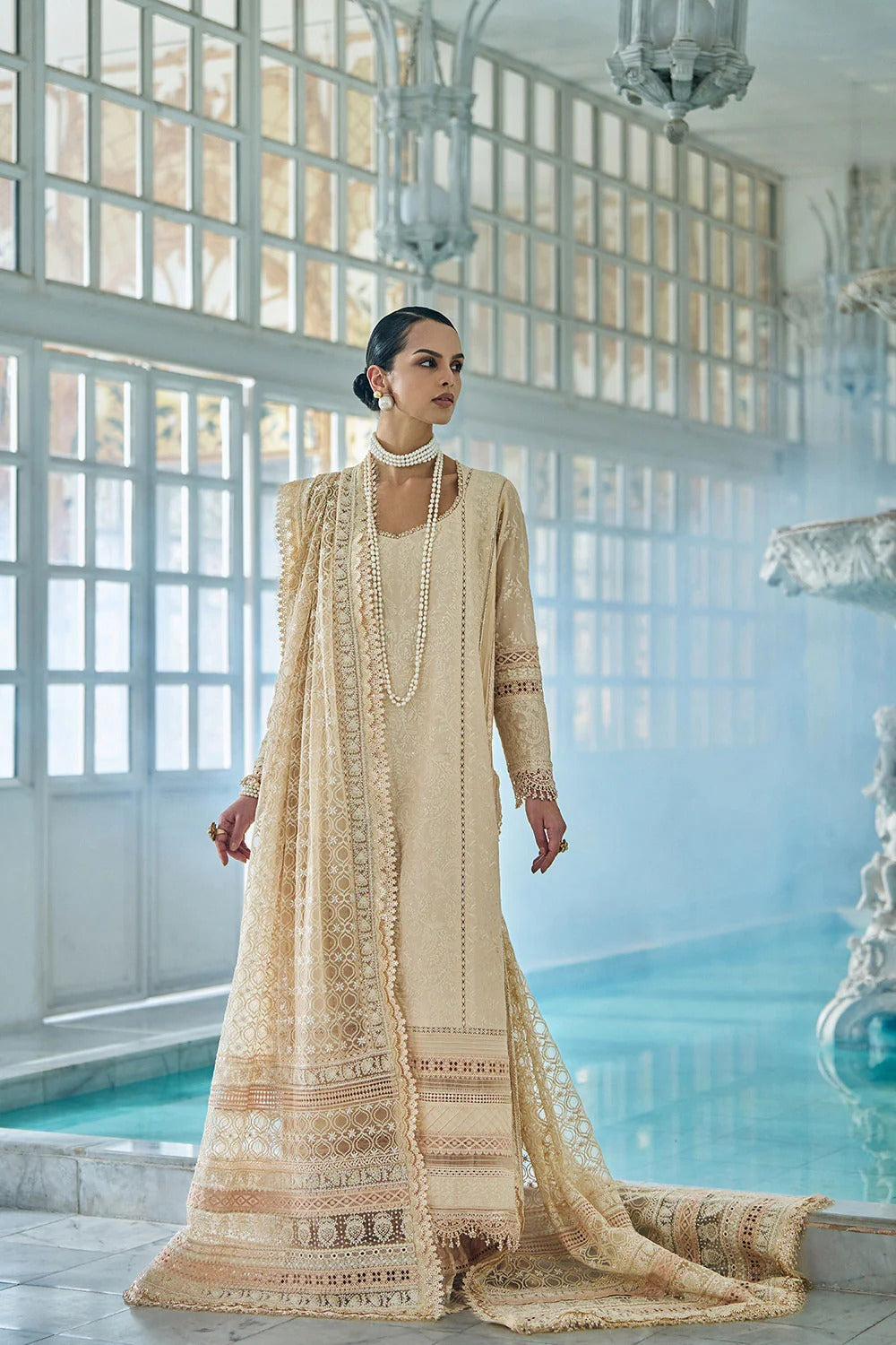 Woman in a traditional beige embroidered outfit standing by a pool with large windows in the background