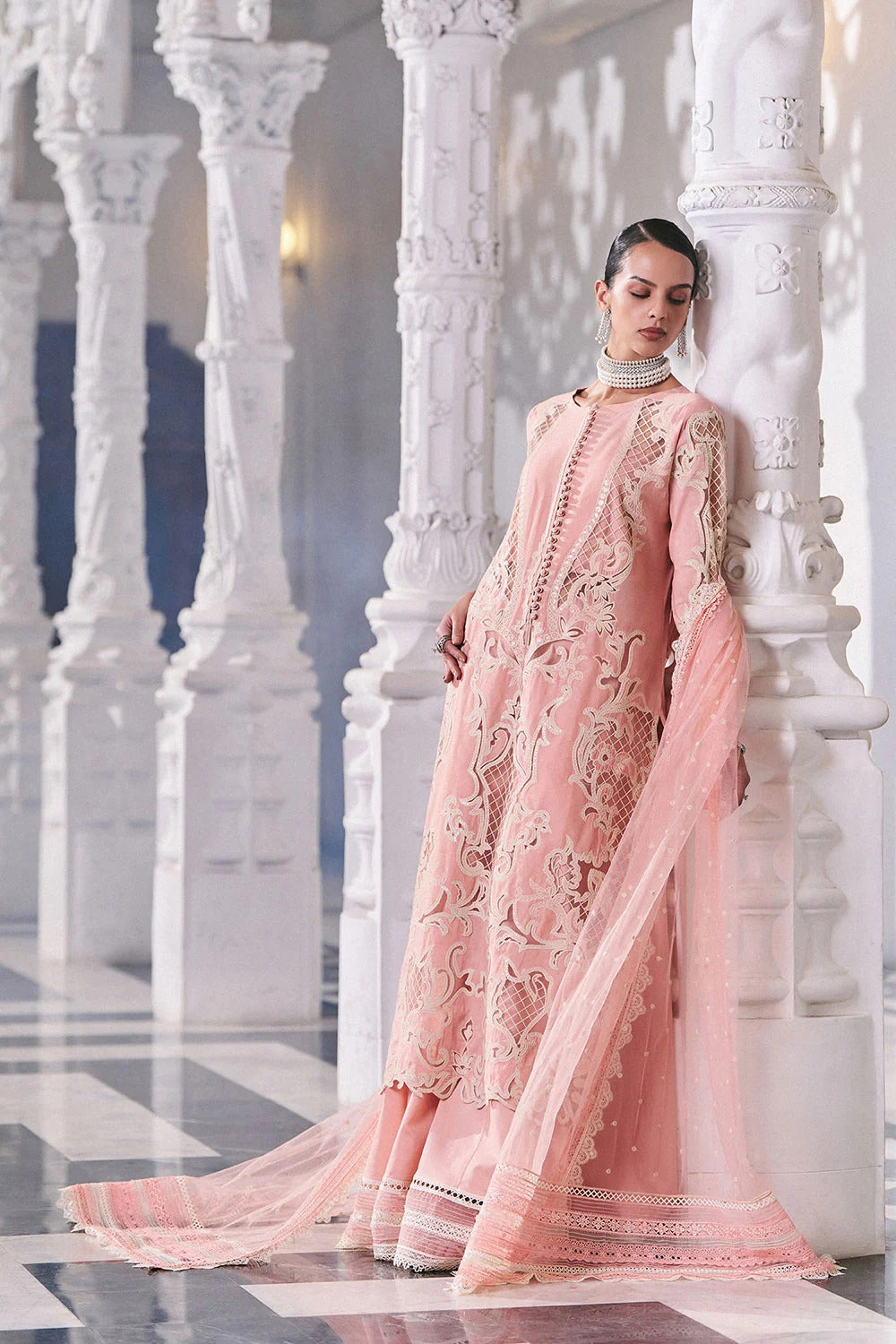 Woman in a pink embroidered outfit standing against white marble columns.