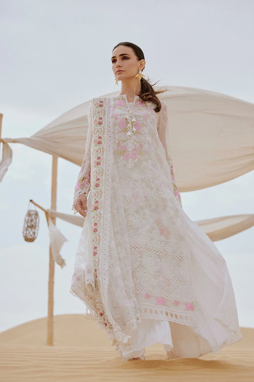 Woman in a white embroidered traditional outfit standing in a desert setting with a tent in the background.
