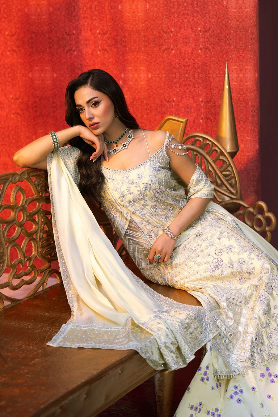 Woman in a traditional outfit sitting on a decorative chair against a red wall.