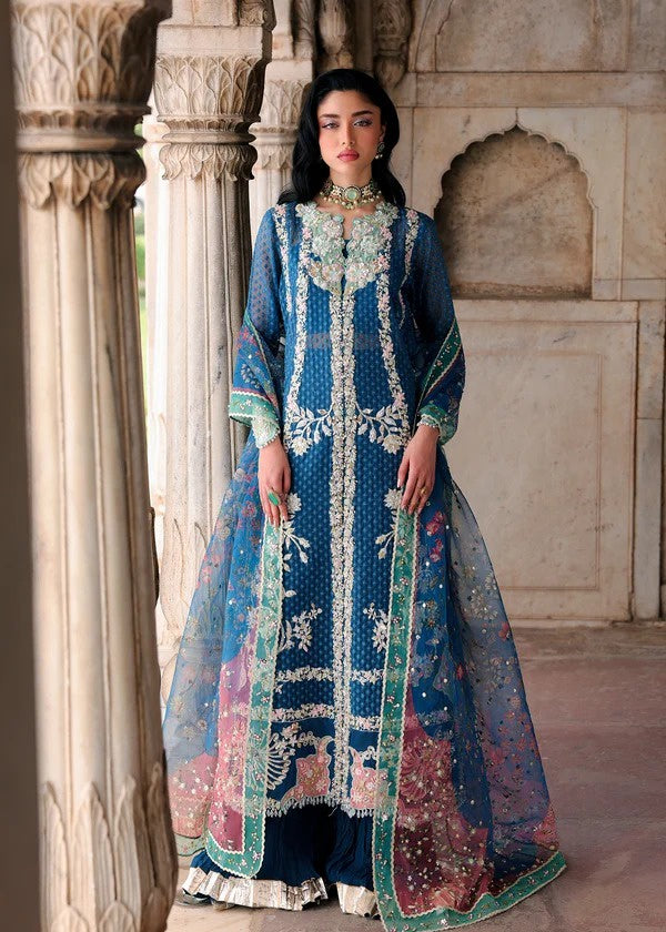 A woman stands in a stone architectural corridor wearing an ornate blue traditional embroidered gown with white and green detailing and a sheer matching dupatta.