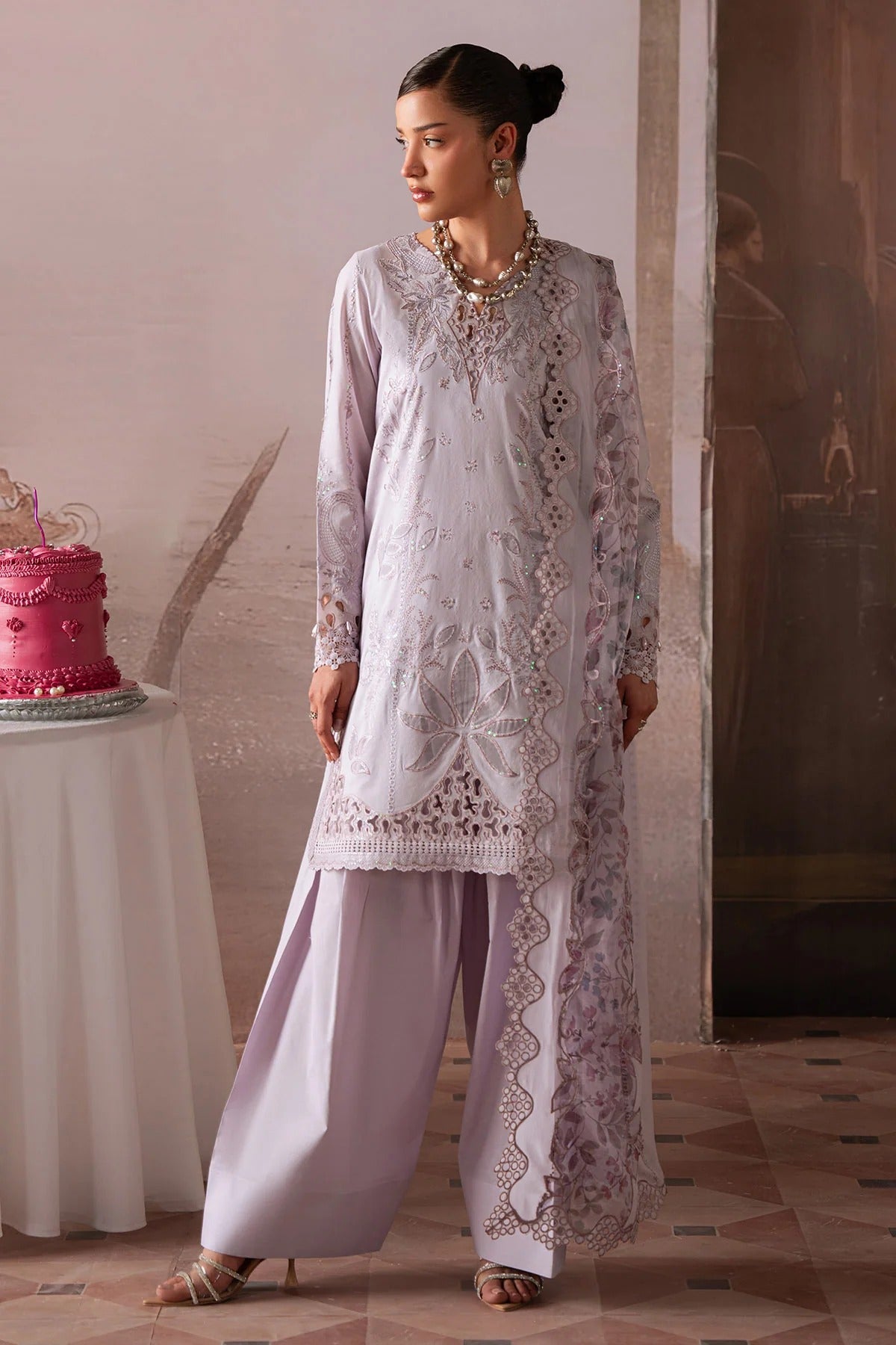 Woman in a light purple embroidered outfit standing next to a pink cake on a table.