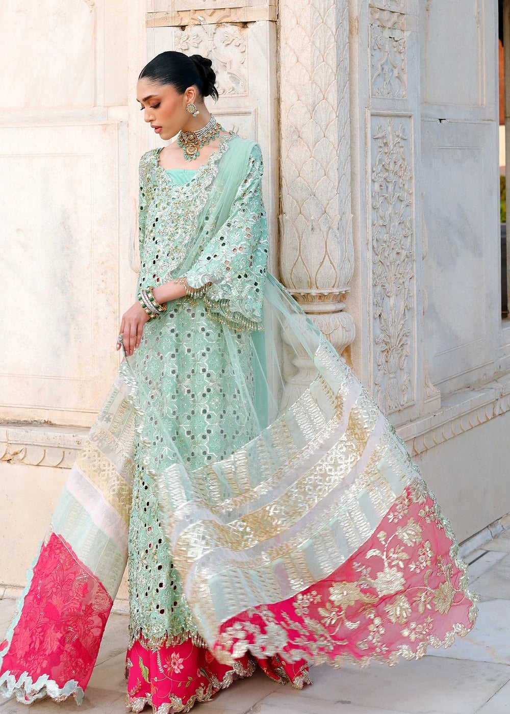 A woman models a traditional mint green embroidered ethnic outfit with a pink dupatta and flared pants, set against an ornate beige stone building.
