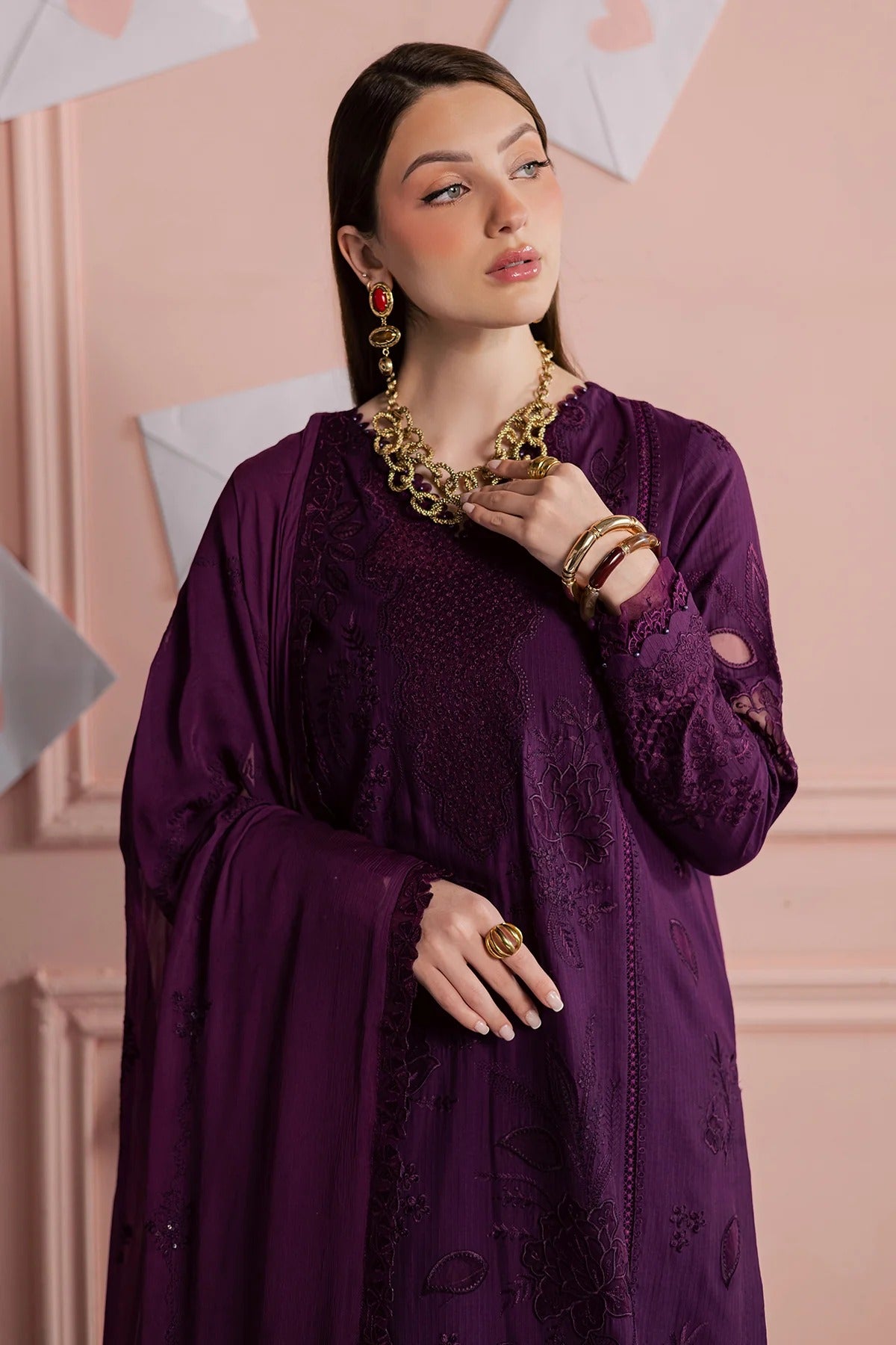 Woman wearing a purple outfit with gold jewelry against a pink wall.