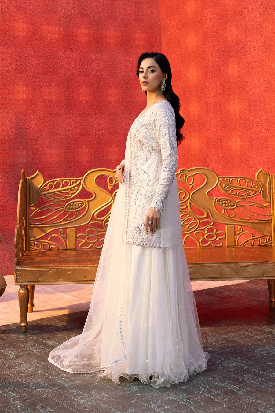 Woman in a white traditional outfit standing in front of an ornate wooden bench with a red patterned wall.