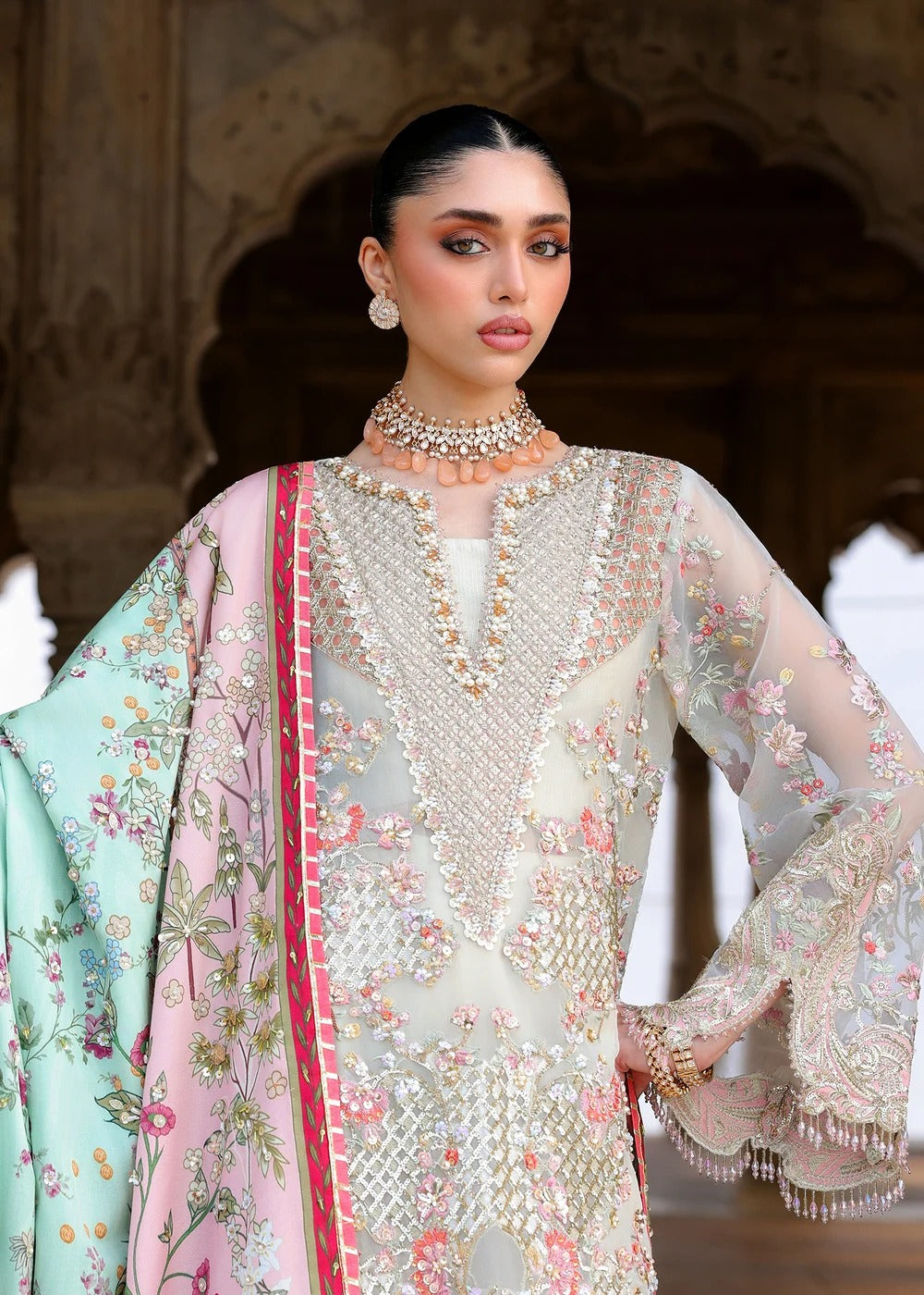 A model wearing a traditional South Asian outfit featuring a heavily embellished cream-colored kurta with floral embroidery, a light pink dupatta with a mint green border and floral print, and a mint green dupatta with a pink border. She is posing in front of an ornate stone archway.
