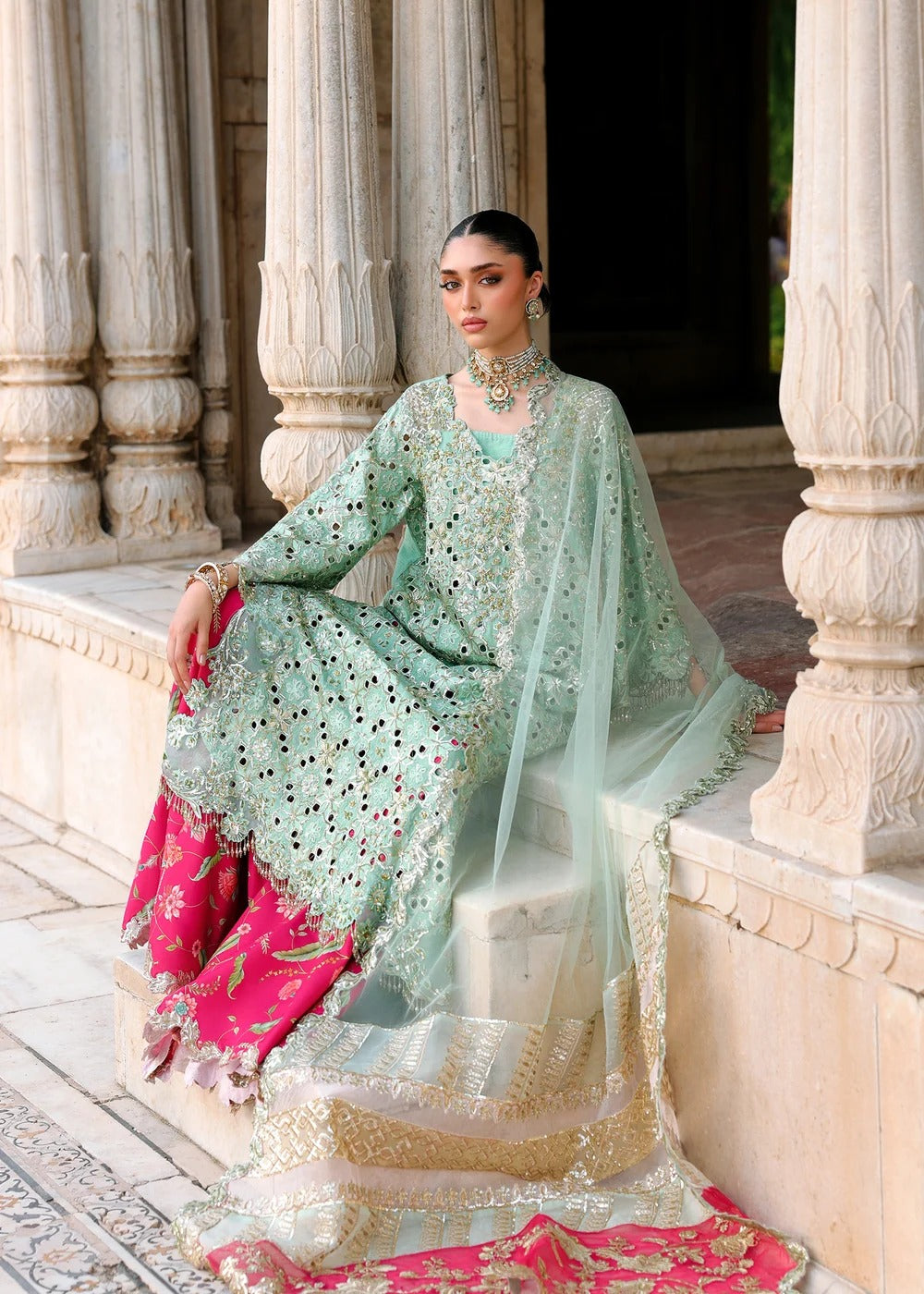 A woman models an ornate mint green embroidered traditional outfit with gold detailing, a matching sheer dupatta, and a bright pink patterned undergarment, posed in front of classical stone pillars.