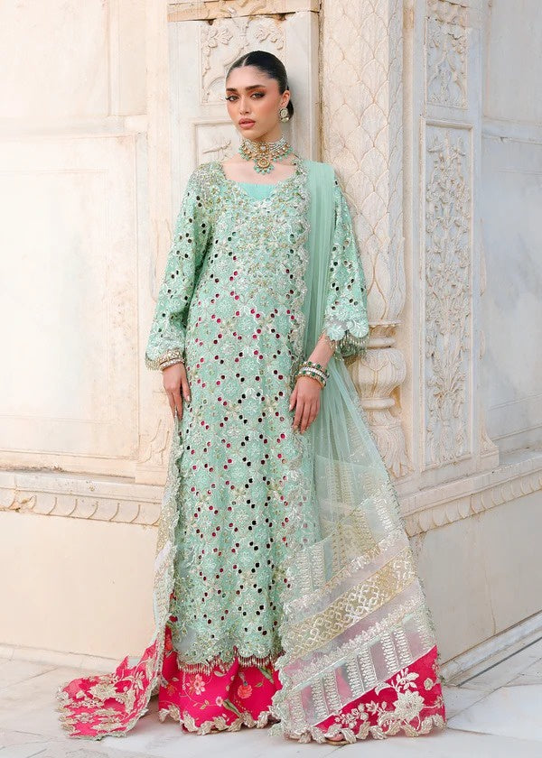 A woman models a mint green and pink heavily embroidered traditional outfit with a matching dupatta, set against a light-colored stone architectural background.