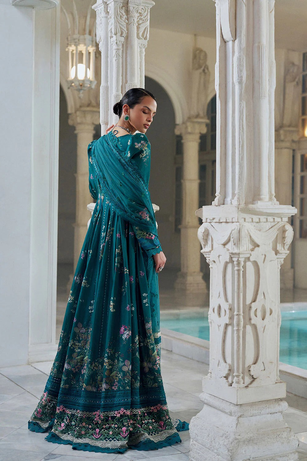 Woman in a teal floral dress standing in an elegant indoor setting with columns and a pool.