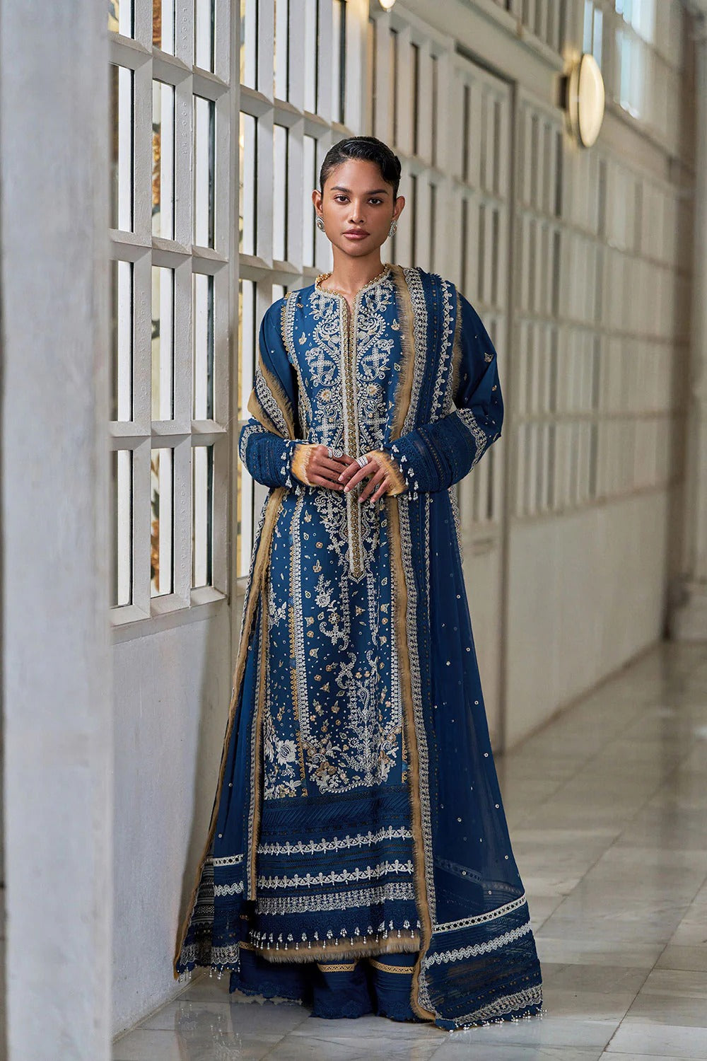 Woman in a blue embroidered traditional outfit standing in a modern indoor setting.
