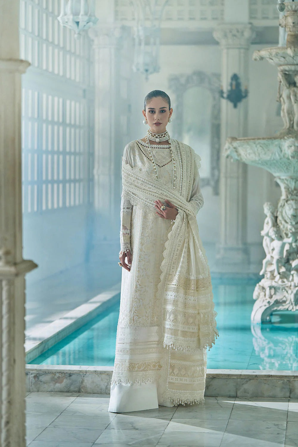 Woman in a white saree standing in a luxurious room with marble pool and columns.