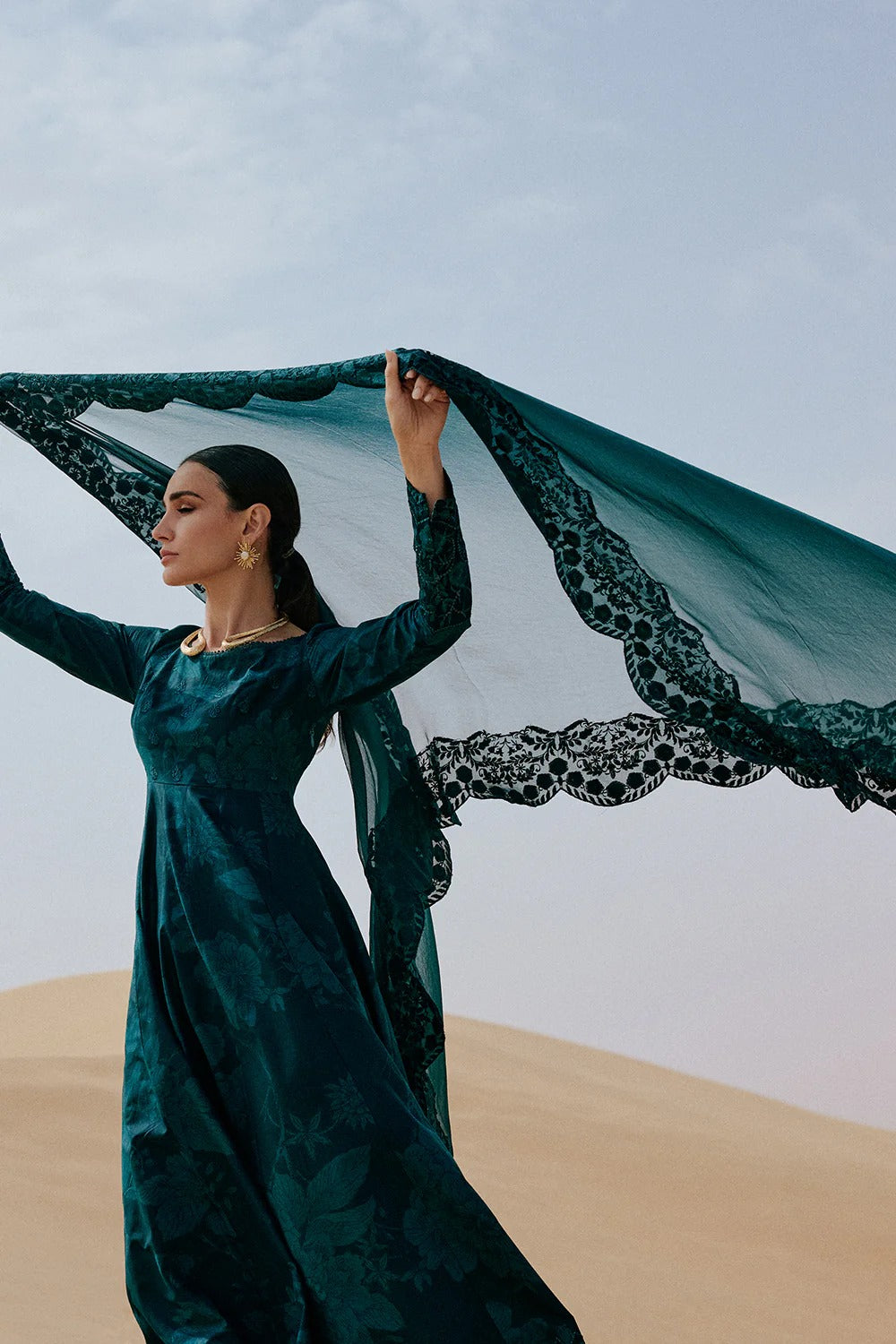 Woman in a green dress holding a black lace scarf against a blue sky and sand dunes.