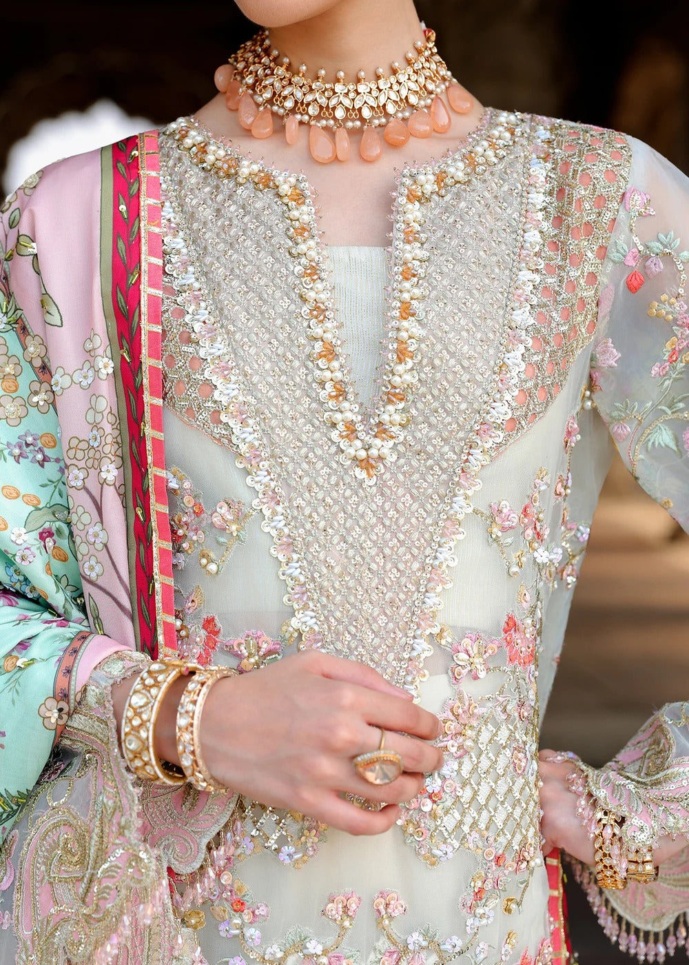 A close-up of a model wearing an ornate traditional outfit with intricate silver, gold, and pink floral embroidery, paired with a pink dupatta and gold jewelry.