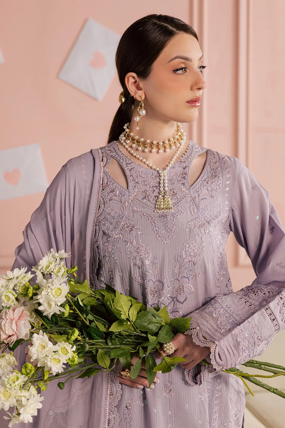 Woman in a light purple embroidered outfit holding flowers against a pink background