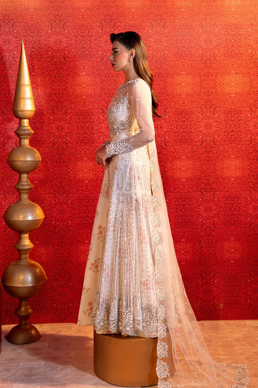 Woman in a white embroidered traditional outfit standing against a red wall with decorative elements.