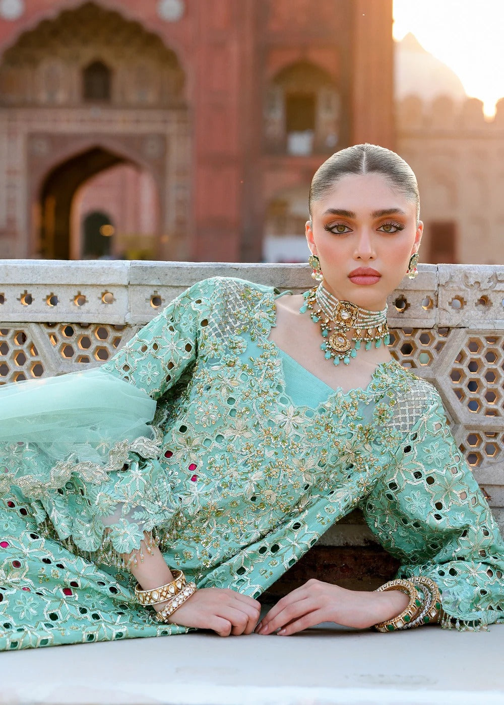 A woman models a mint green embroidered traditional outfit with gold mirror work and turquoise accents, posed in front of a historic red sandstone building.