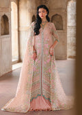 A woman models a heavily embroidered peach-pink traditional gown with a matching sheer dupatta in a historic stone corridor.