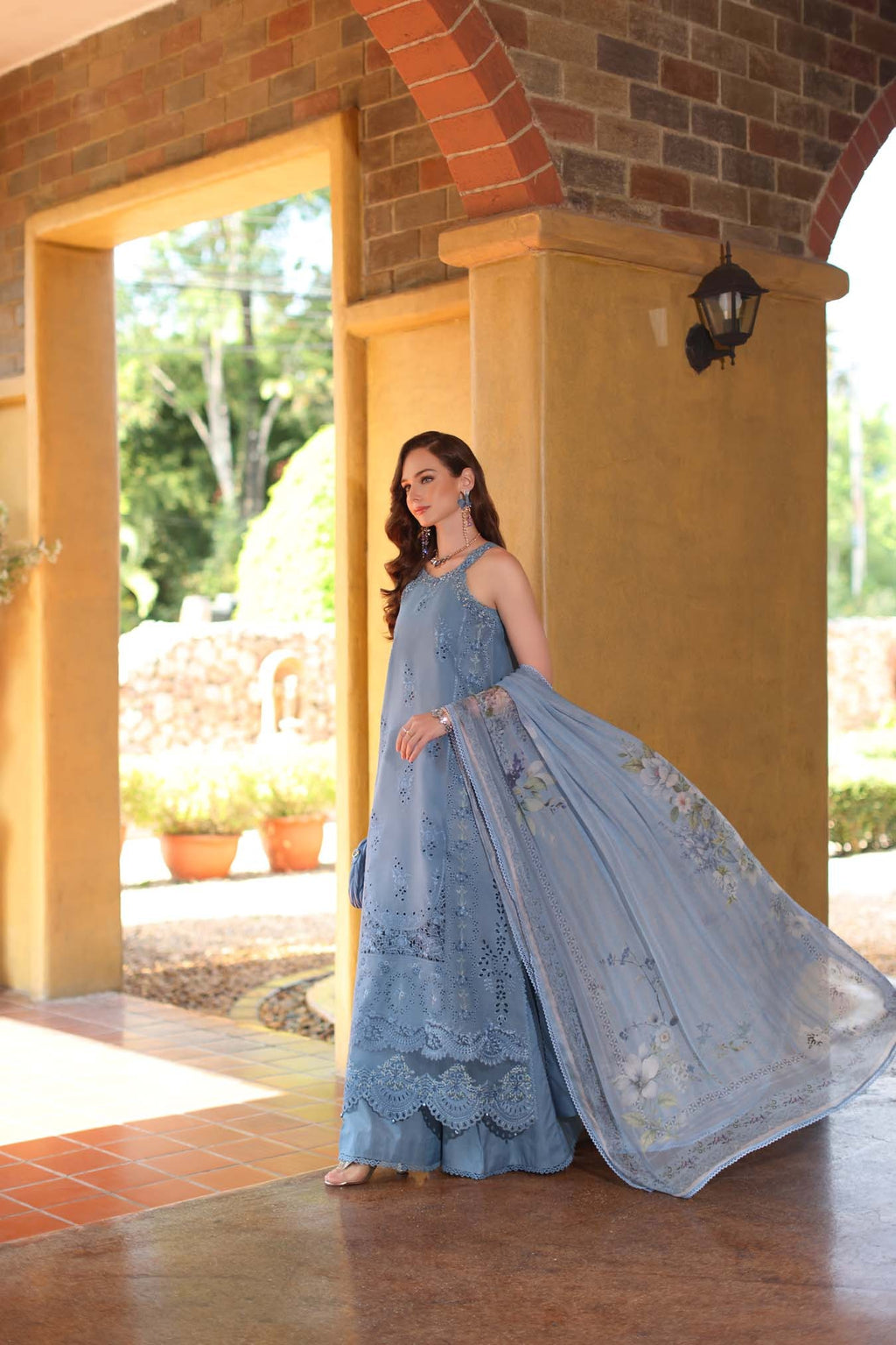 Woman in a light blue embroidered dress standing in front of a brick wall with an archway.