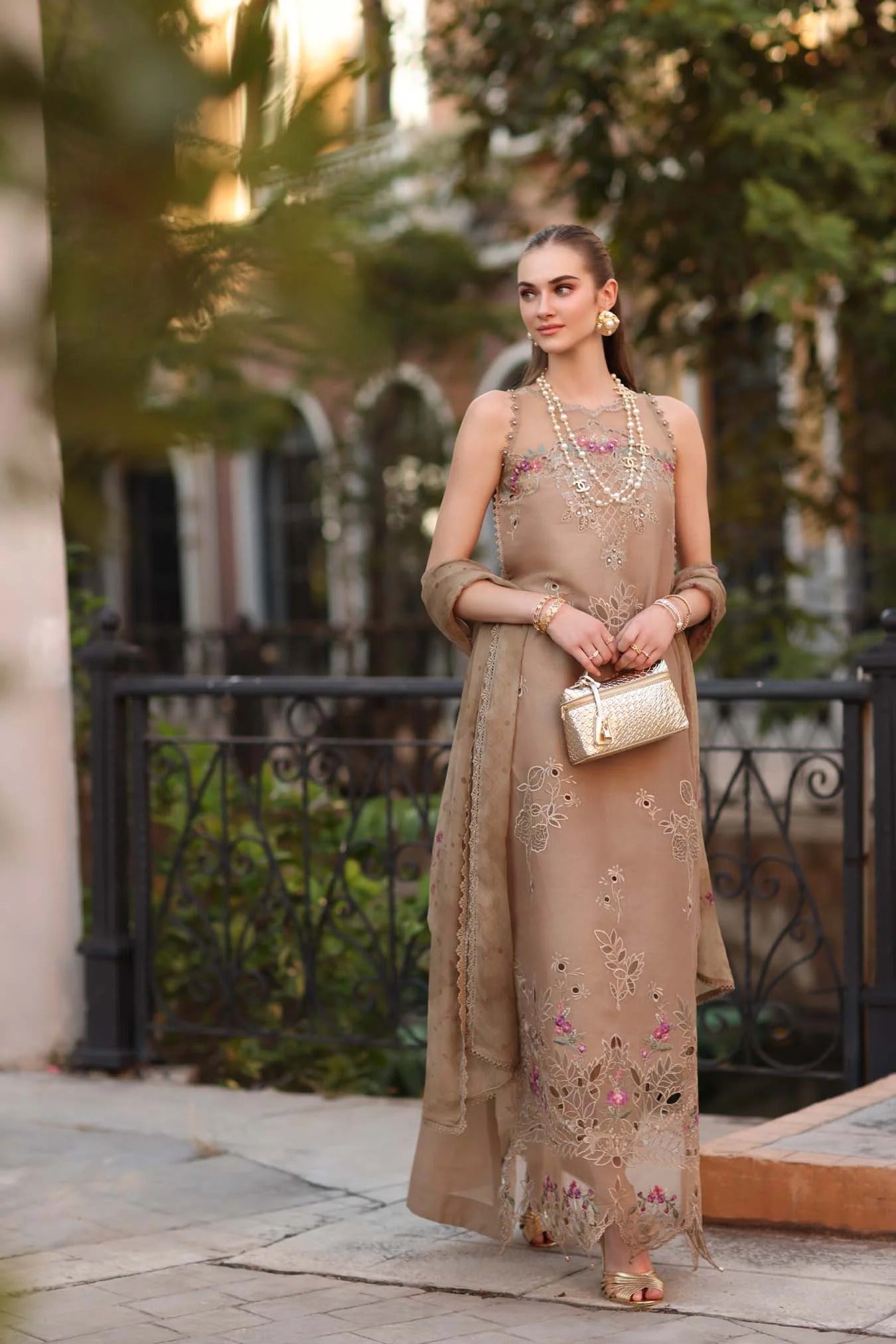 Woman in a beige embroidered dress standing outdoors with greenery in the background