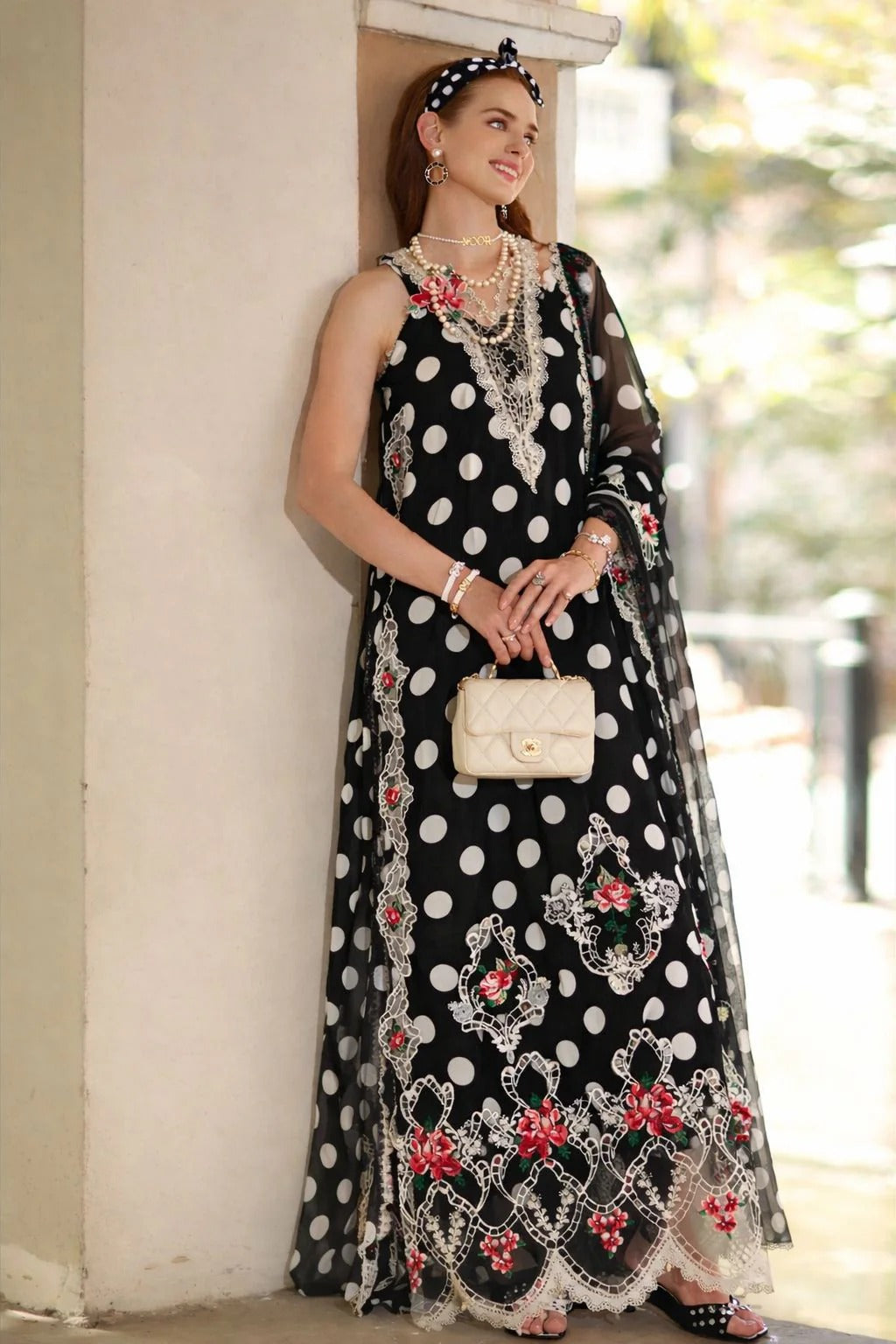 Woman wearing a black polka dot dress with floral patterns, standing against a blurred outdoor background.