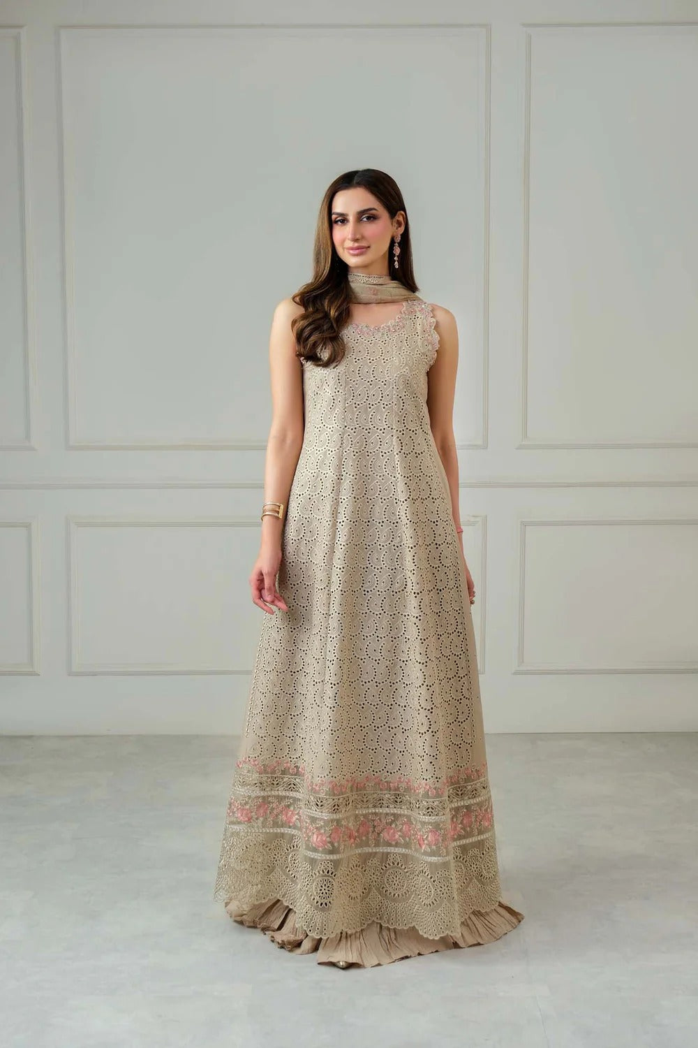 Woman wearing a long beige dress with floral patterns against a white paneled wall.