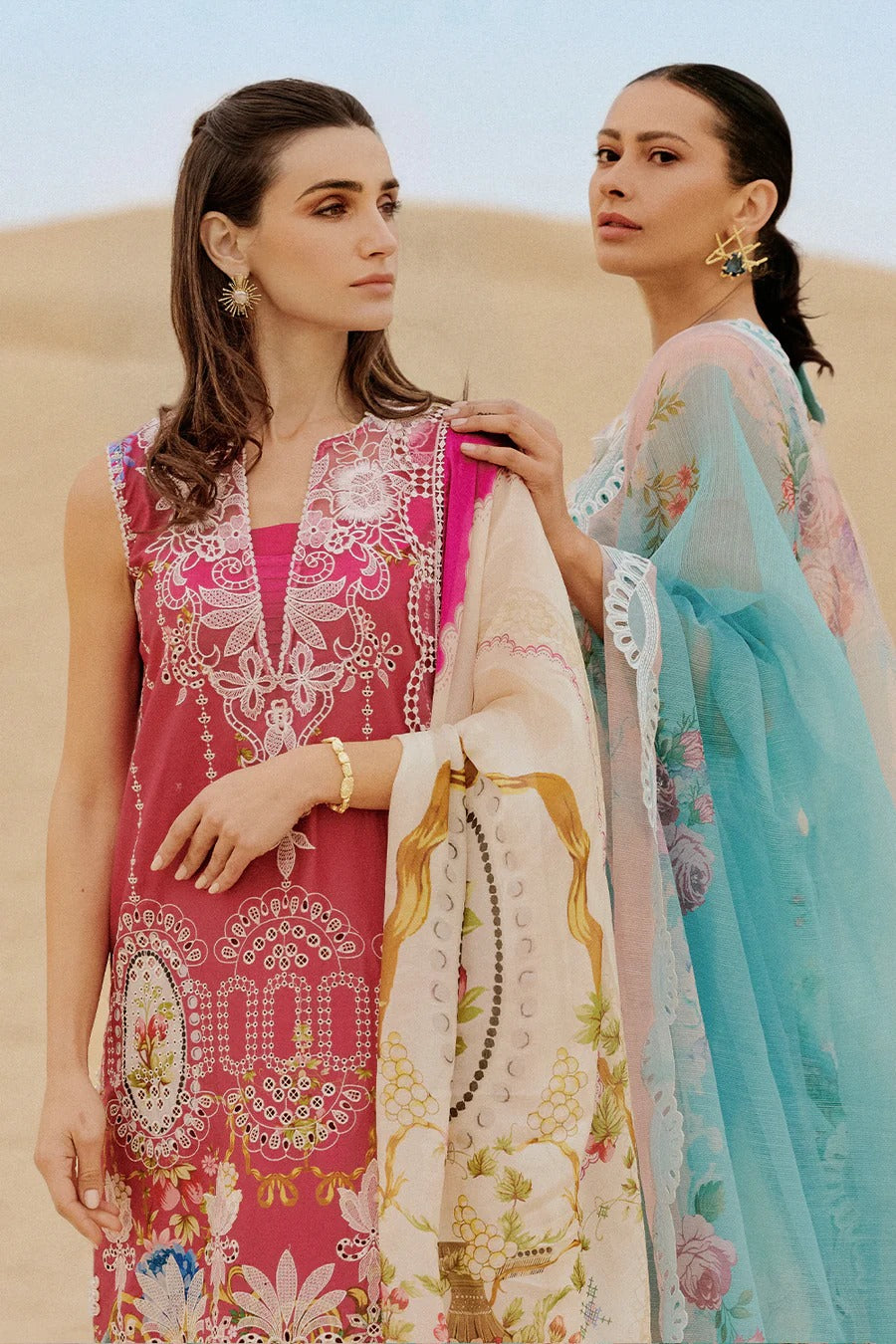 Two women in traditional embroidered dresses standing in a desert setting.