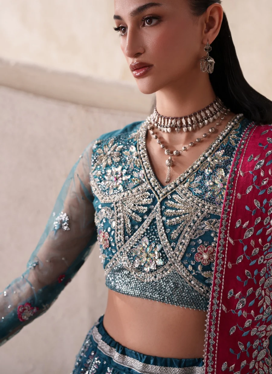 A close-up of a model wearing an intricately embroidered teal blue and pink traditional Indian outfit with silver mirror work, layered jewelry, and a red dupatta with floral embroidery.