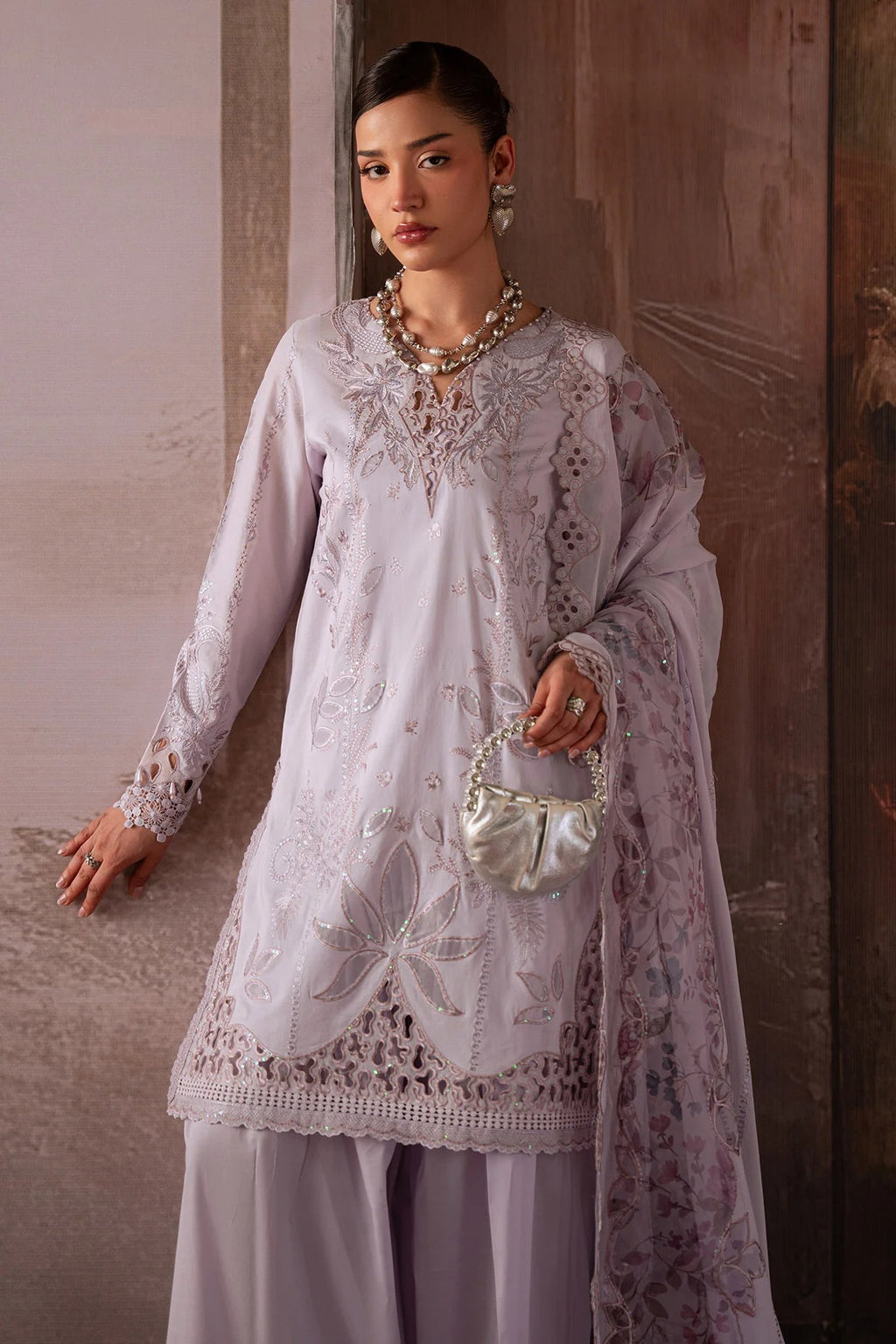 Woman wearing a white embroidered outfit with a necklace and handbag against a neutral background