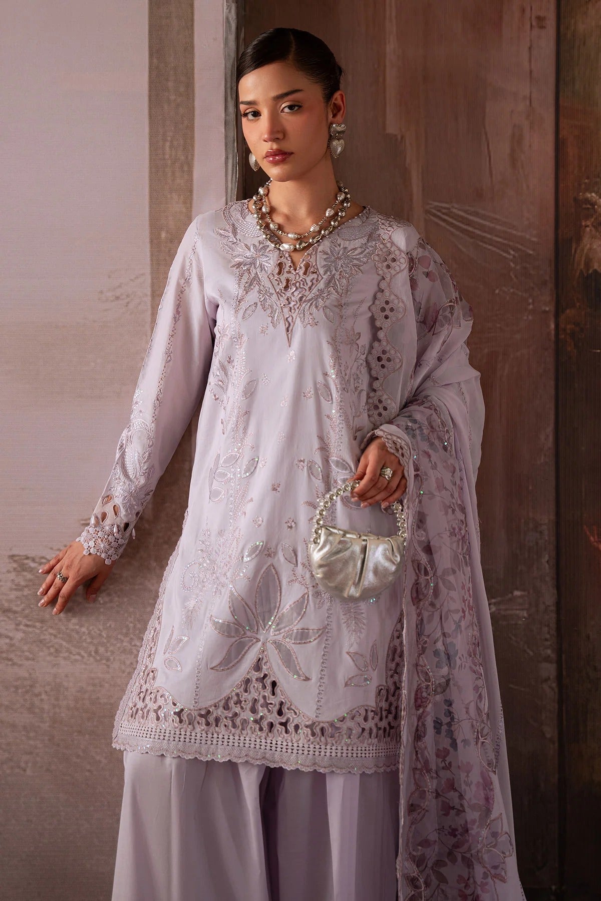 Woman wearing a white embroidered outfit with a necklace and handbag against a neutral background