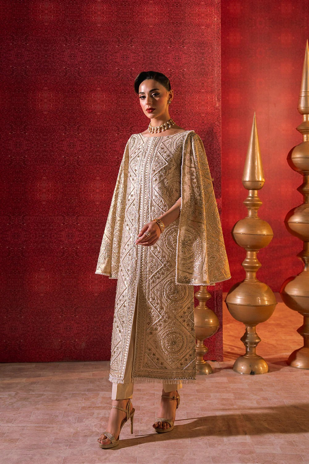 Woman in a cream embroidered outfit standing against a red wall with decorative elements.