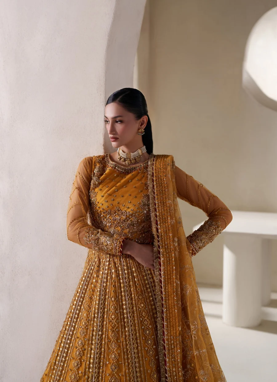 A woman modeling a traditional Indian ensemble consisting of a mustard yellow embroidered lehenga, a matching embroidered blouse, and a sheer dupatta, set against a minimalist beige architectural background.