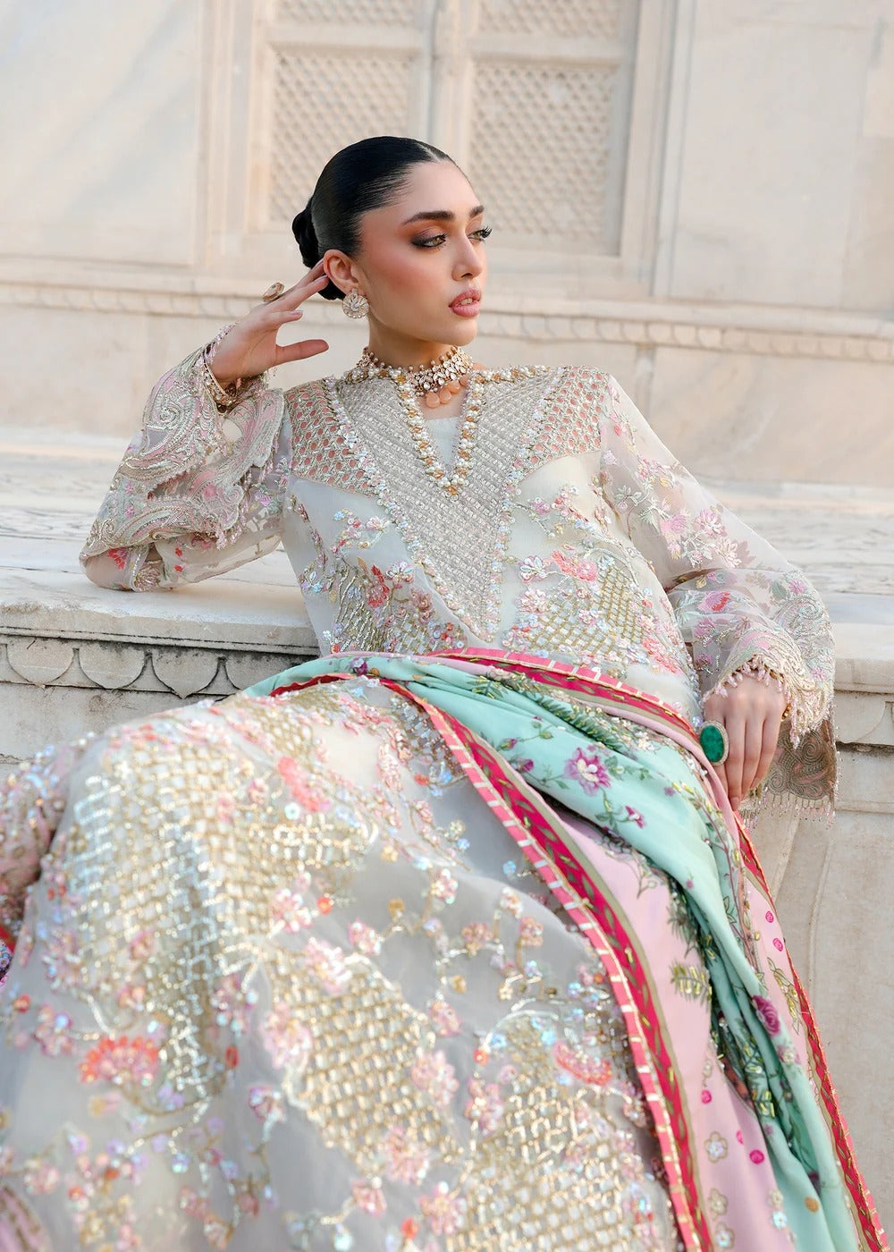 A woman models a traditional South Asian outfit featuring a heavily embroidered light gray and pink gown with a mint green dupatta accented with red borders, set against a classical stone background.