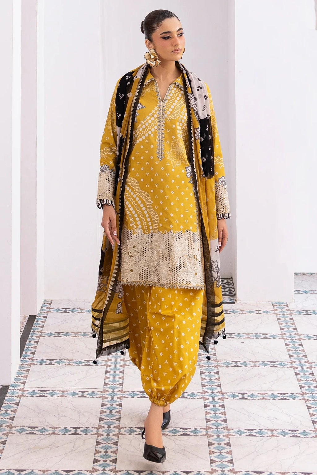 Woman in a yellow traditional outfit with a black dupatta in a room with patterned tiles.