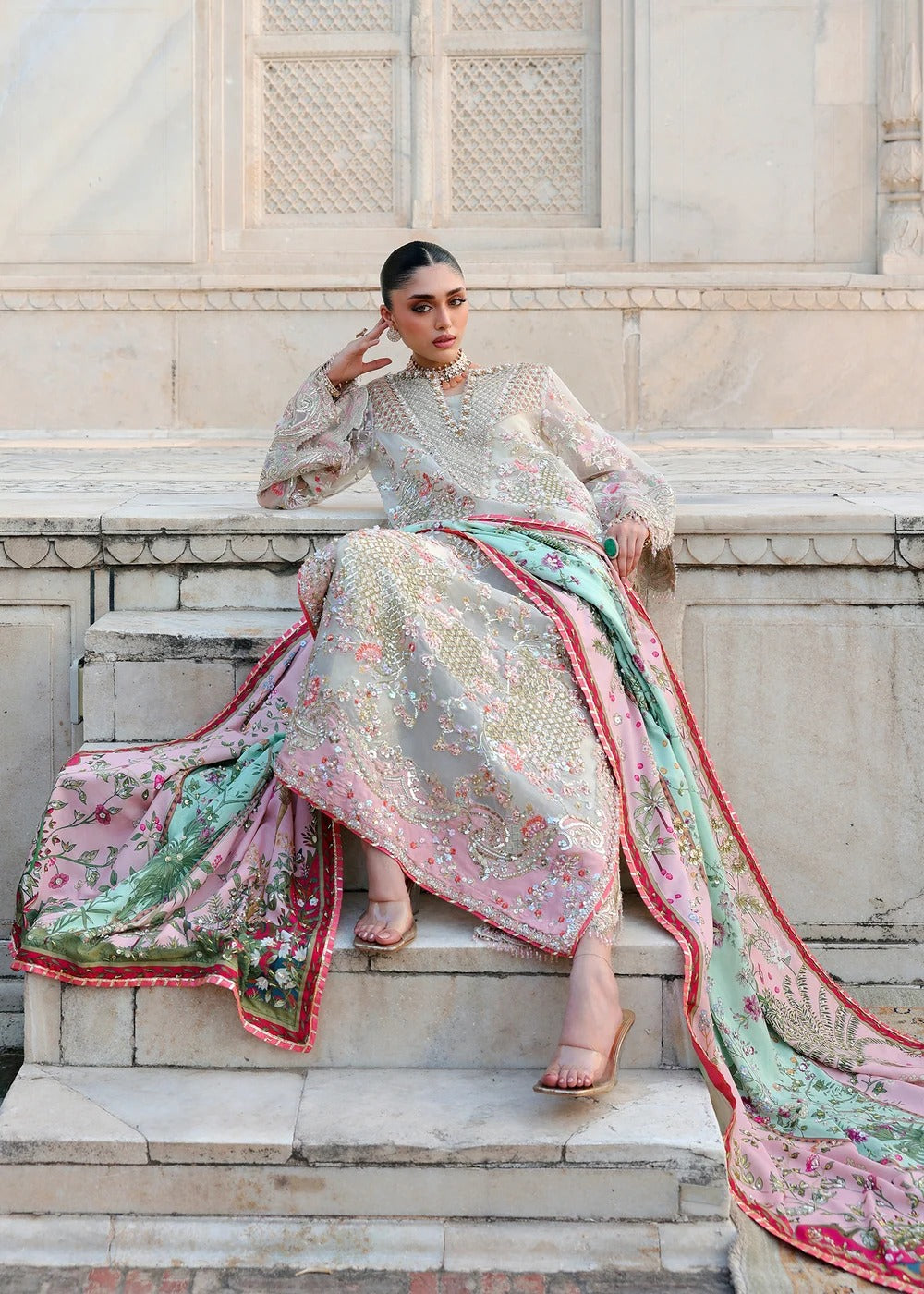 A woman models a traditional South Asian outfit consisting of a heavily embroidered light beige kurta and a matching long dupatta with pink, green, and red floral patterns, seated on stone steps.