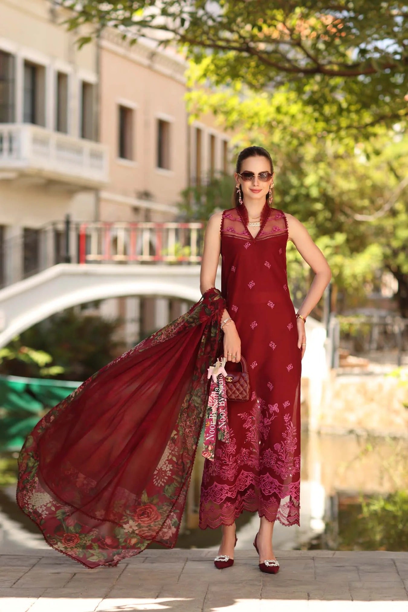 Woman in a red dress with a long scarf standing outdoors near a bridge.