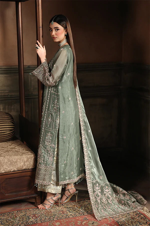 Woman in a traditional green embroidered outfit standing in a room with wooden furniture.