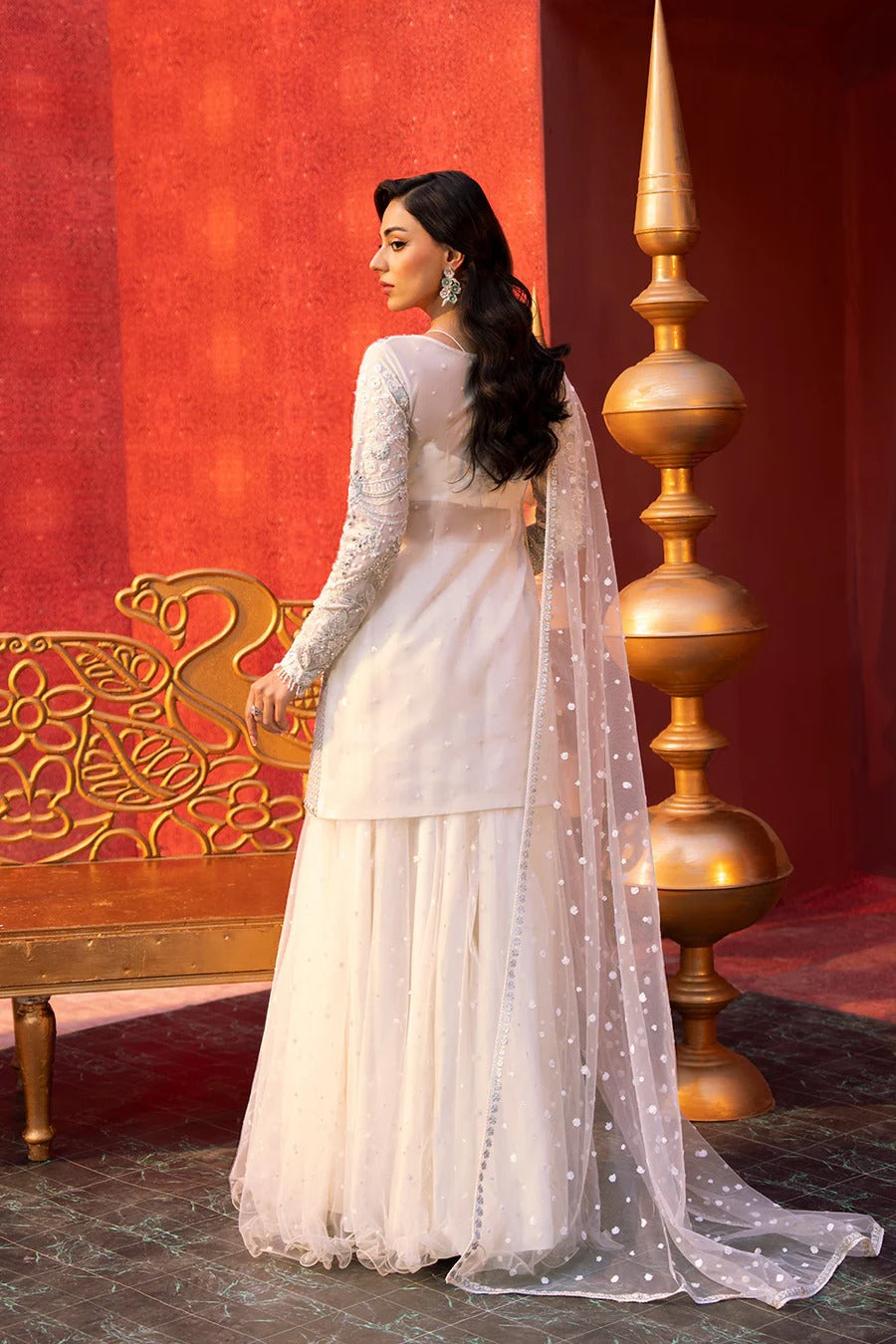 Woman in a white traditional outfit standing in front of a red wall with gold decorative elements.