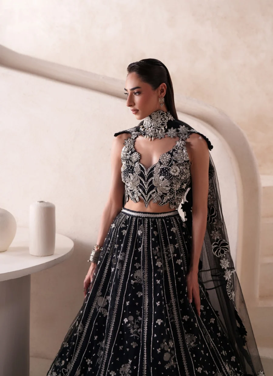 A woman models a black and silver heavily embellished Indian ethnic wear set, featuring a high-neck embroidered crop top, a matching flared skirt with intricate silver mirror work, and a sheer black dupatta draped over her shoulders, set against a warm beige architectural background.