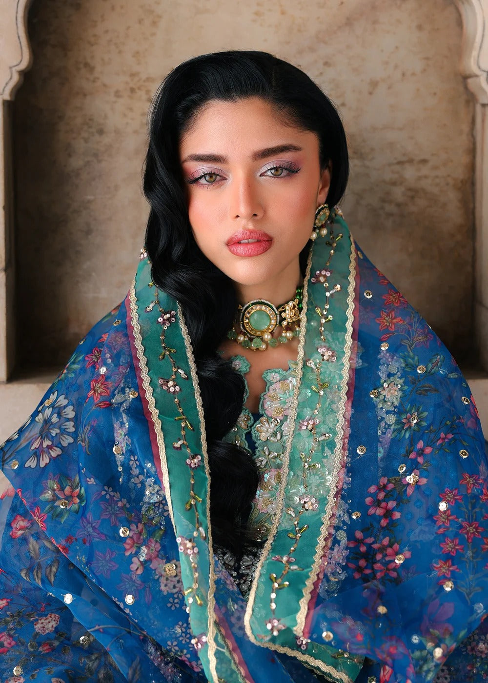 A woman wearing traditional South Asian attire with a rich blue and teal embroidered dupatta and matching embellished top, styled with a statement gold necklace, set against a textured beige stone background.