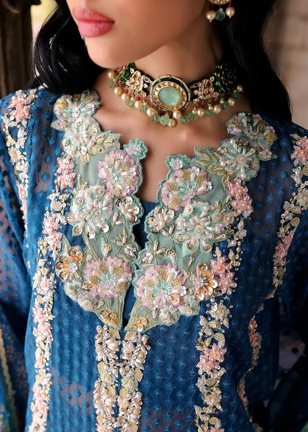 A close-up of a model wearing a detailed blue traditional embroidered garment with floral embroidery in pink, green, and gold, paired with a matching ornate choker necklace.