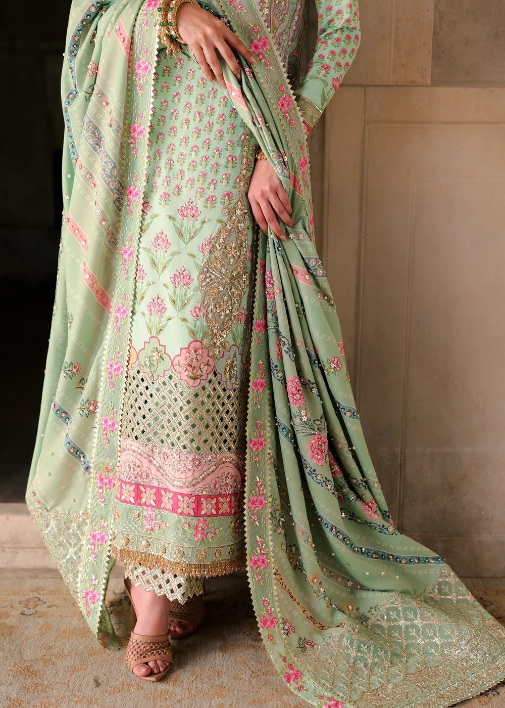 A sage green ethnic salwar suit with pink and gold floral embroidery, shown on a model. The set includes a kurta, matching trousers, and a draped dupatta, photographed against a neutral stone wall background.
